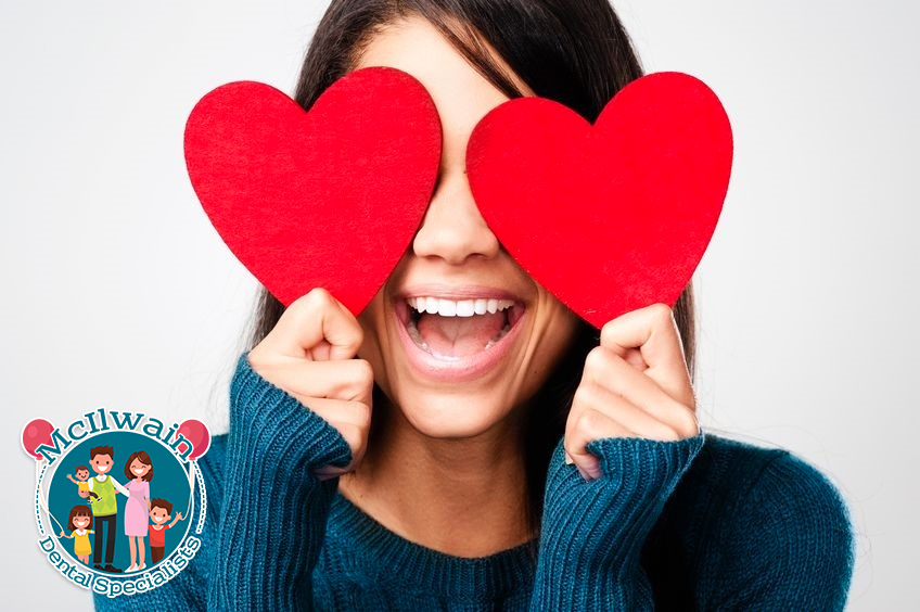 Woman holding two red hearts over her eyes next to a logo of McIlwain Dental Specialists.