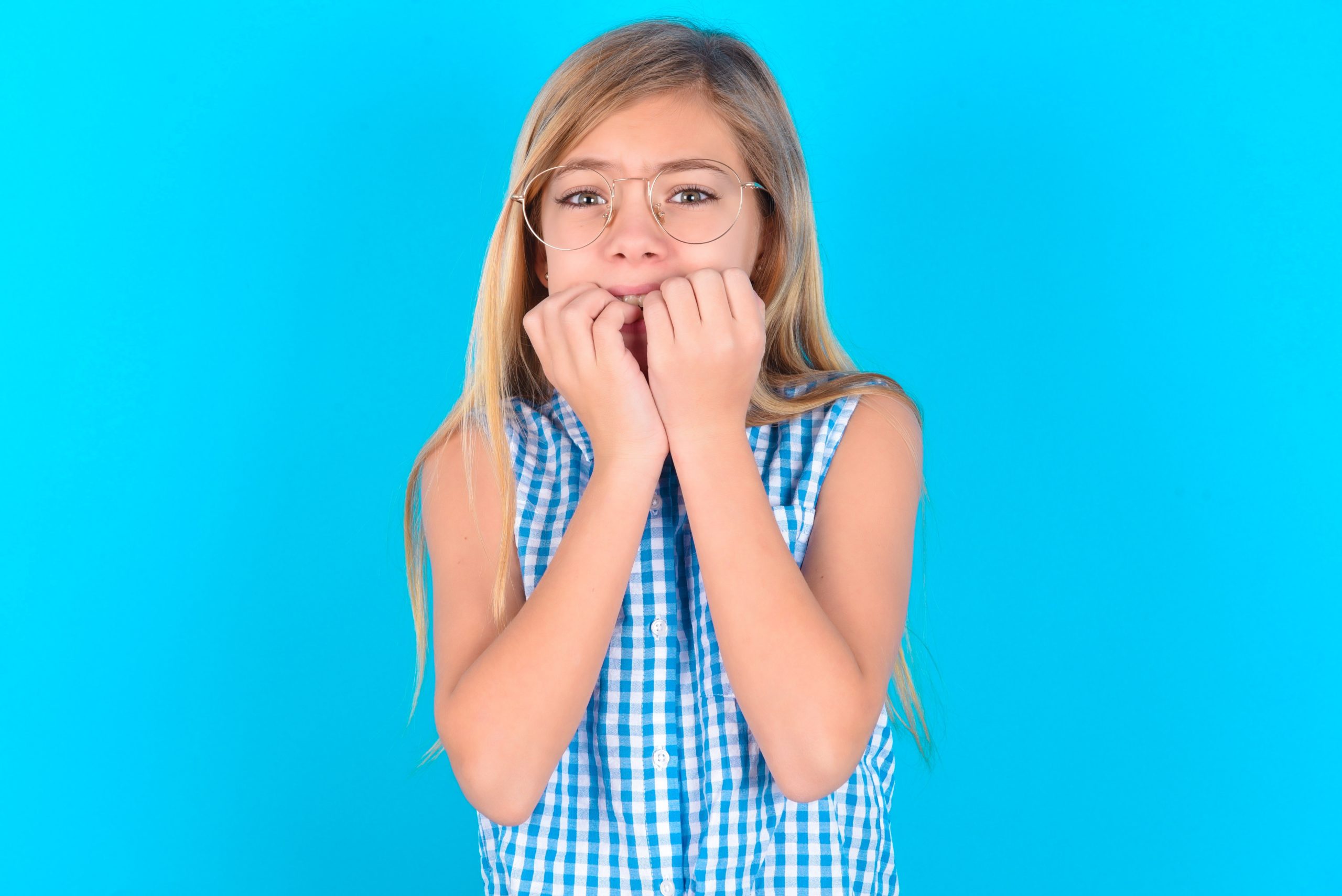 A young blonde girl holds her hands in front of her mouth in apprehension. She stands in front of a light blue background