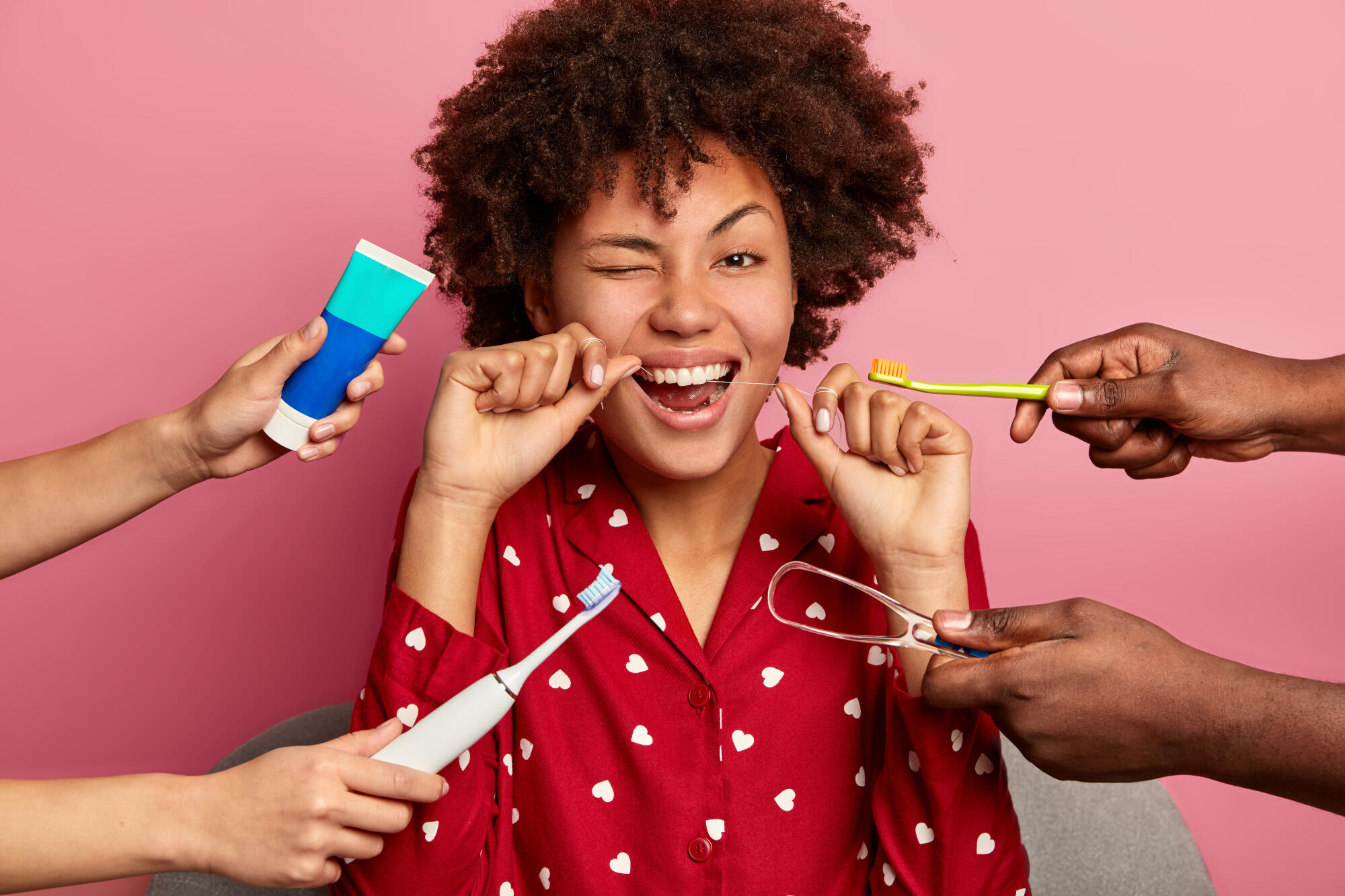 Person flossing in a red heart-patterned shirt with multiple hands offering dental care products against a pink background.