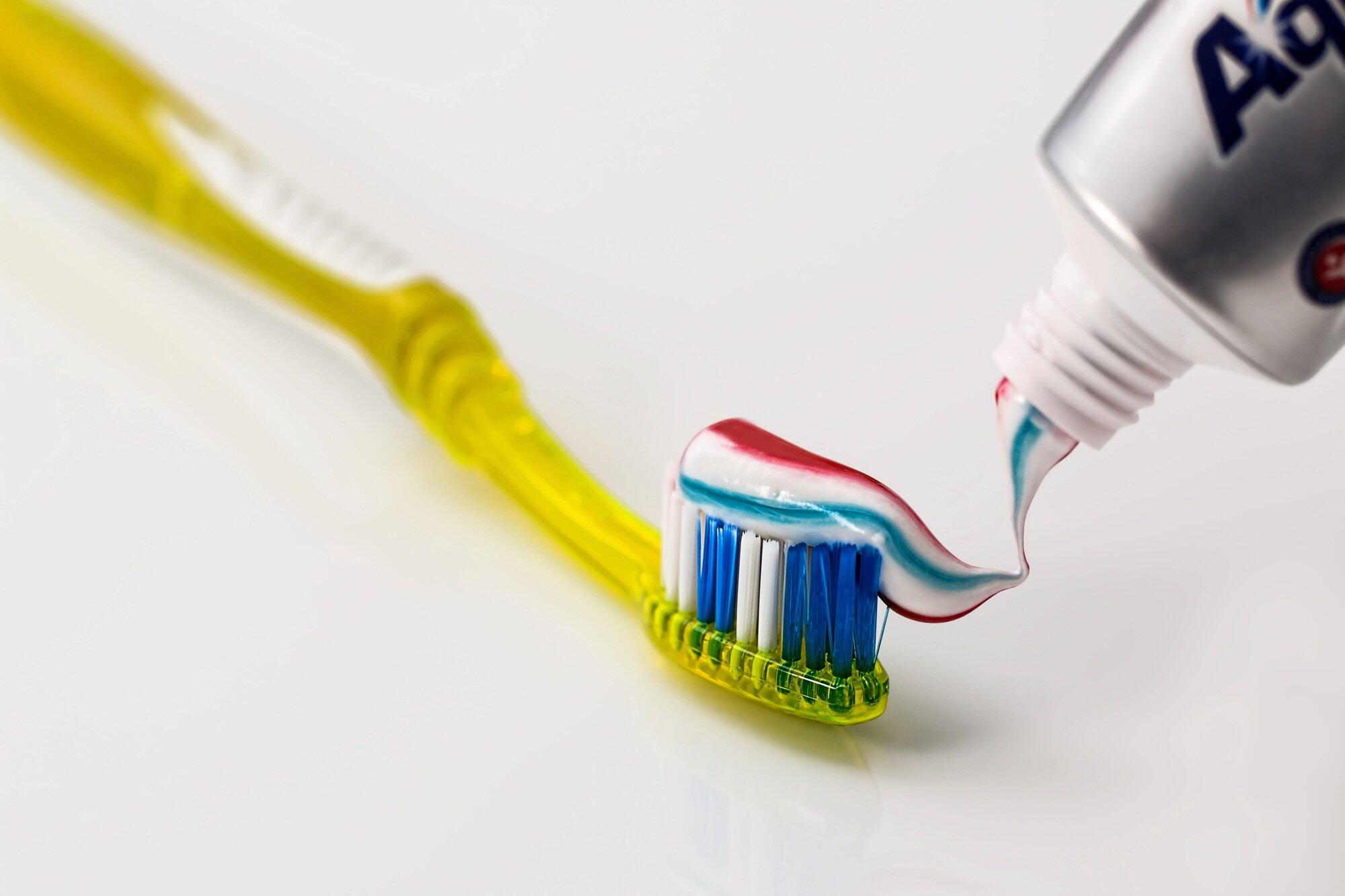 A yellow toothbrush with blue bristles with red and blue striped toothpaste being squeezed from a tube.